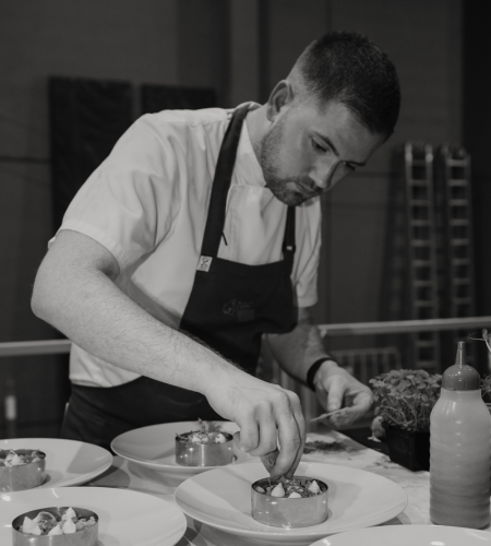 A black and white image of QPAC's Executive Sous Chef, James Gallagher. James wears an apron and is placing a garnish on the plates in front of him.